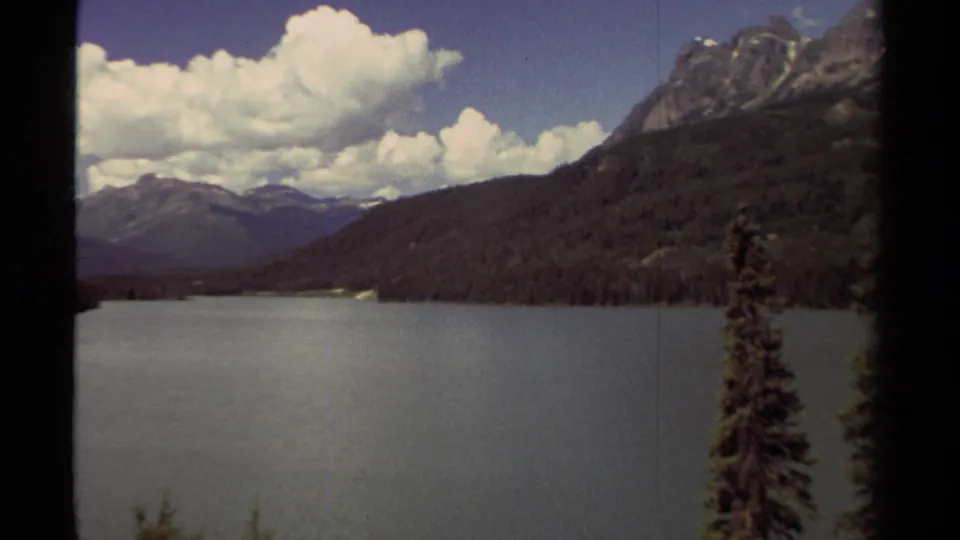 1979 British Columbia lake scenery with evergreen-covered mountains, distant ridges, bright clouds, and a foreground conifer.