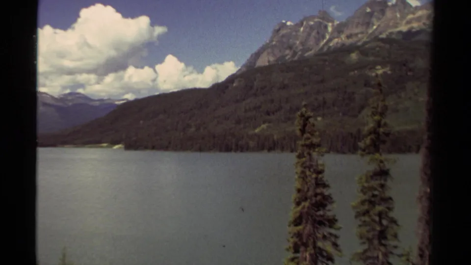 1979 8mm British Columbia mountain lake view with steep wooded slopes, foreground conifers, and scattered white clouds.
