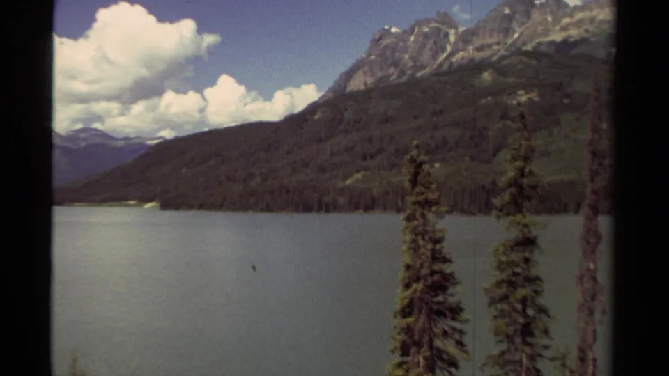 1979 British Columbia home-movie frame showing a mountain lake, evergreen foreground trees, wooded slopes, and distant ridges.