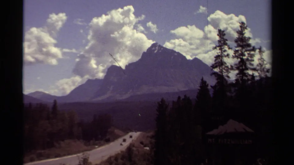 Soft 8mm frame of a British Columbia mountain road, forested slopes, distant vehicles, and a Mt. Fitzwilliam sign in shadow.