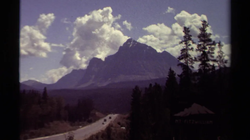 1979 8mm British Columbia mountain-road view with evergreen silhouettes, a distant roadway, cars, clouds, and a shadowed Mt. Fitzwilliam sign.