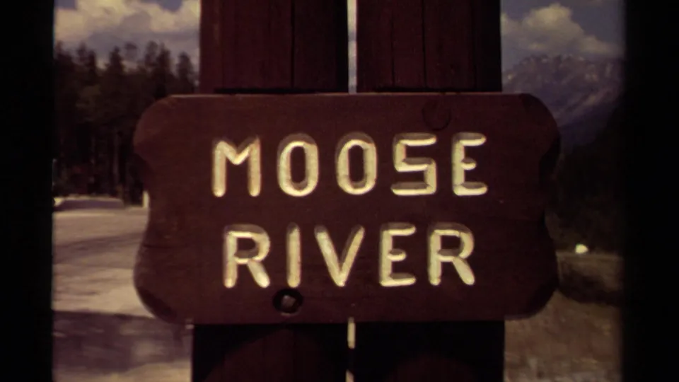 1979 British Columbia 8mm frame showing a carved Moose River sign in front of snowy forest and distant mountains.