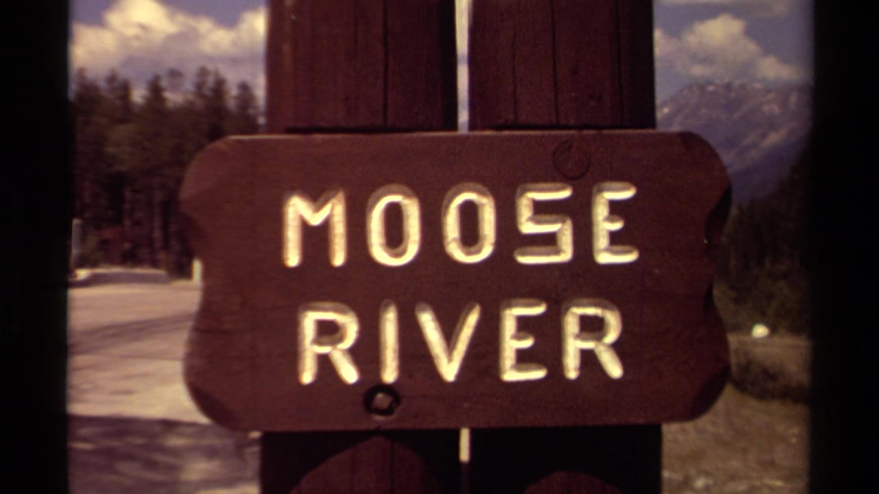 Close 8mm view of the Moose River sign in British Columbia, with snowy ground, trees, and mountain scenery behind it.