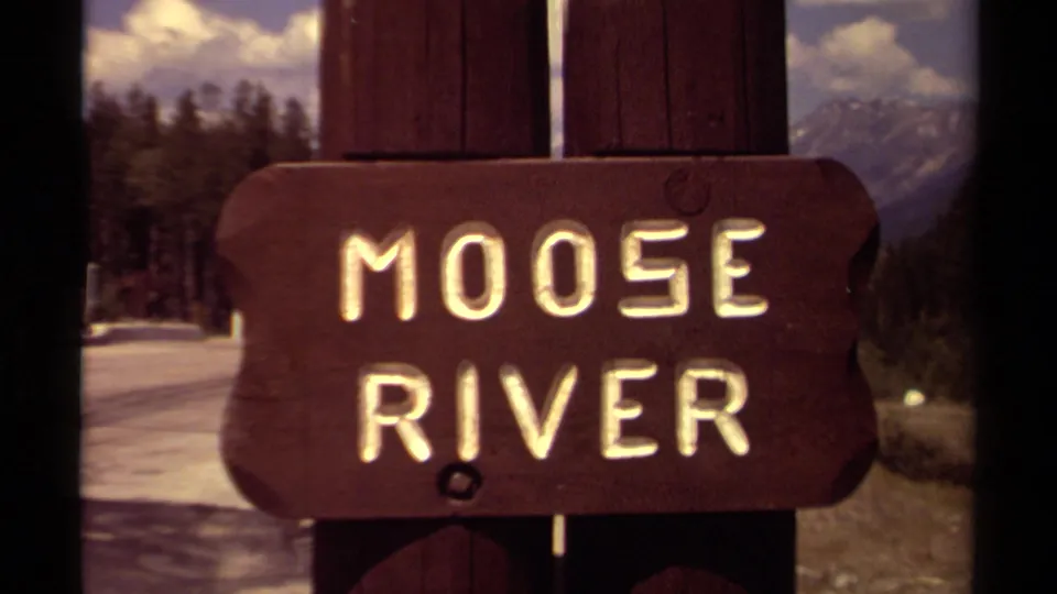 Close 8mm view of the Moose River sign in British Columbia, with snowy ground, trees, and mountain scenery behind it.