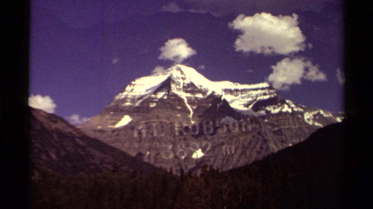 Snow-capped mountain peak rising above dark valley slopes under scattered clouds.