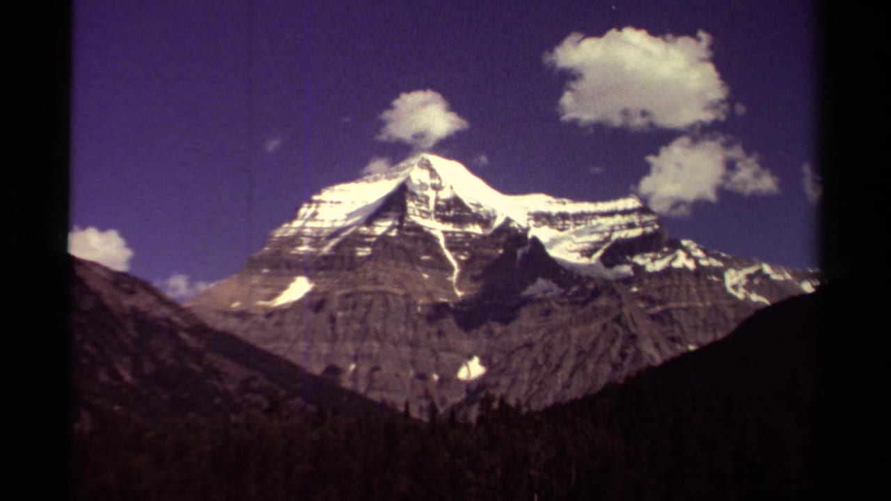 1979 8mm British Columbia mountain view with a snowy peak, rocky slopes, dark foreground hills, and scattered clouds in a purple-blue sky.