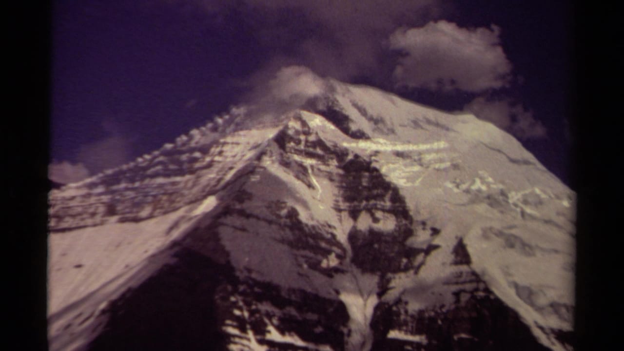 Closer 8mm frame of a British Columbia snow-covered mountain face, with dark rock bands and clouds near the upper ridge.