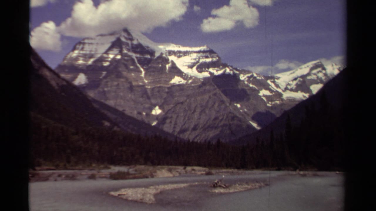Snow-capped mountain range above a river valley and evergreen forest.