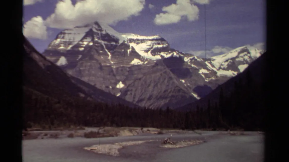 British Columbia 8mm travel footage frames snow-streaked mountains above dark forest and a pale waterway.