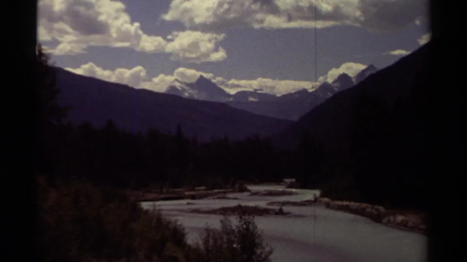 1979 8mm British Columbia frame of a river winding through a forested valley beneath distant snow-bright mountains and clouds.
