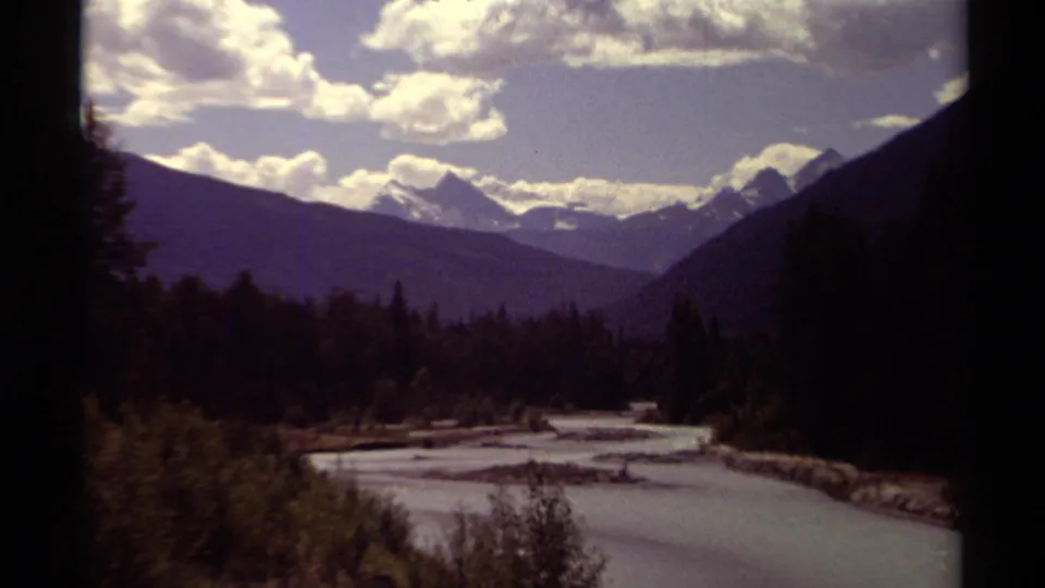 Vintage 1979 8mm view in British Columbia showing a broad river bend, evergreen banks, layered mountains, and a clouded sky.