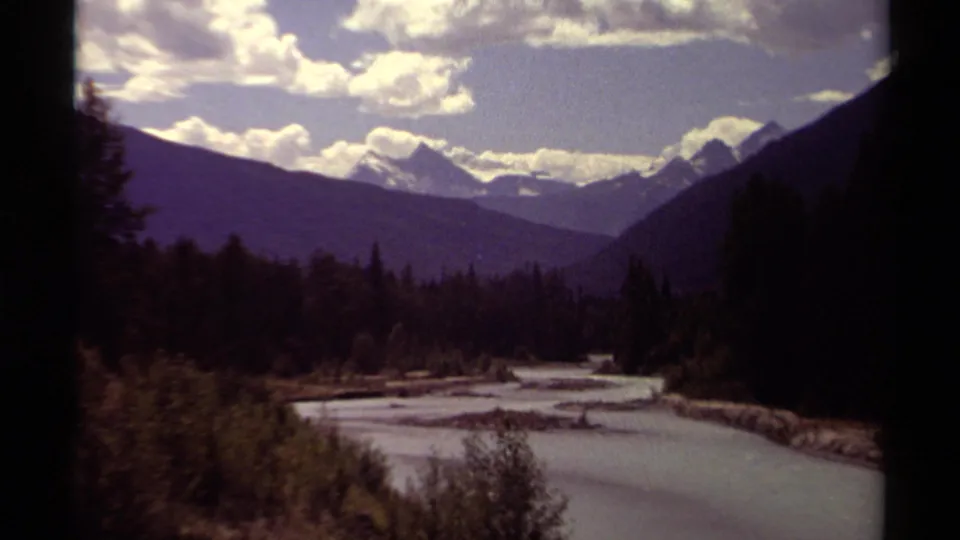 1979 8mm British Columbia home-movie frame with a forest-lined river, purple-toned mountain layers, snowy peaks, and bright clouds.