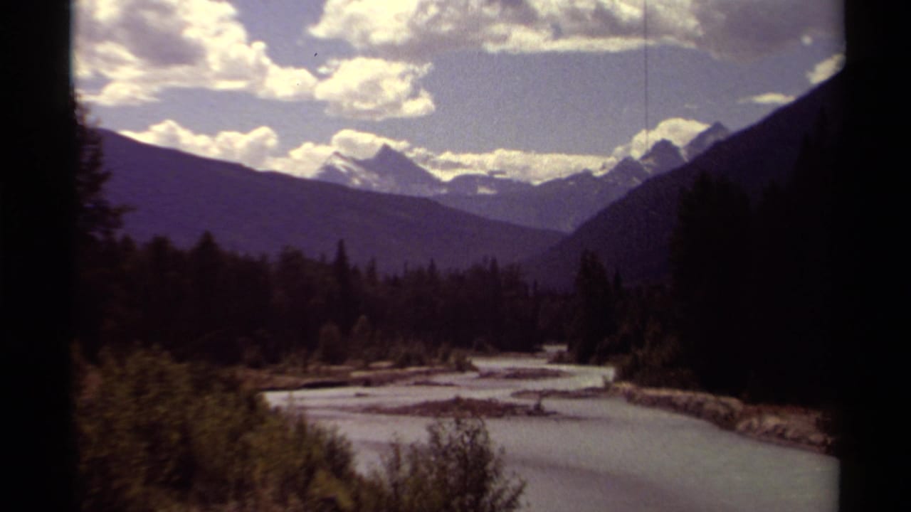 British Columbia mountain valley seen on 8mm film, with a winding river, forested slopes, clouds, and distant snow-covered peaks.