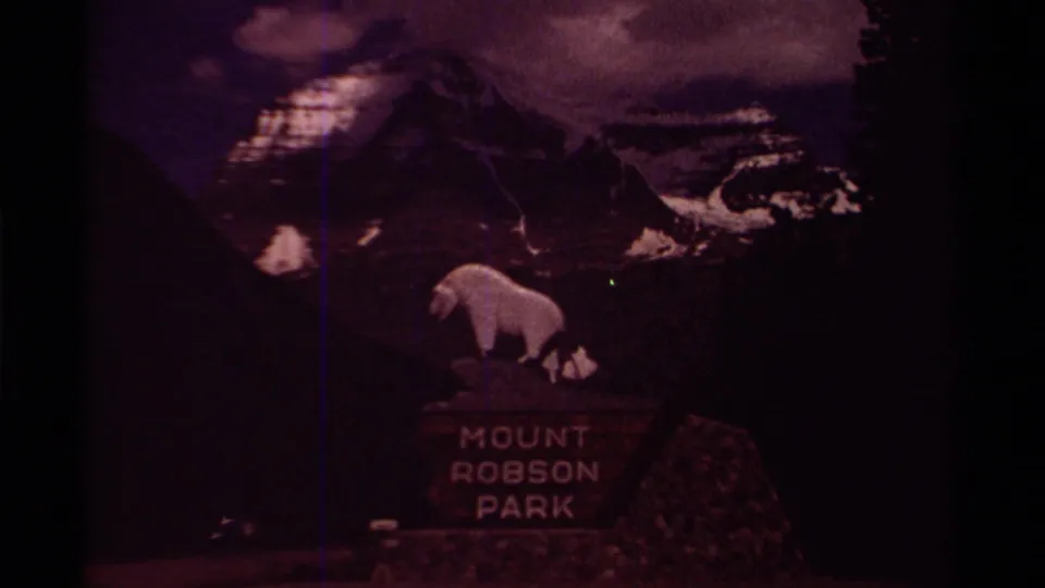 Mount Robson Park entrance sign appears with a white sheep statue, stone base, roadside shadows, and snow-streaked mountain behind it.
