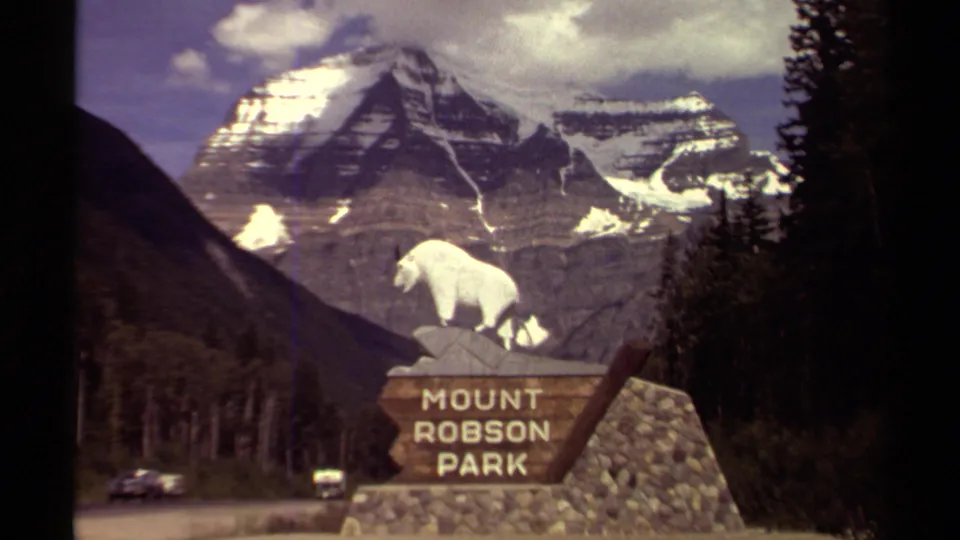 Roadside view of the Mount Robson Park sign in British Columbia, with white sheep statue, rocky base, mountain backdrop, and small cars passing below.