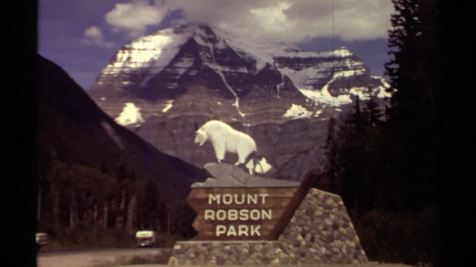 Mount Robson Park roadside sign fills the frame, with a white sheep statue, rocky base, dark tree line, and snow-covered mountain in the background.