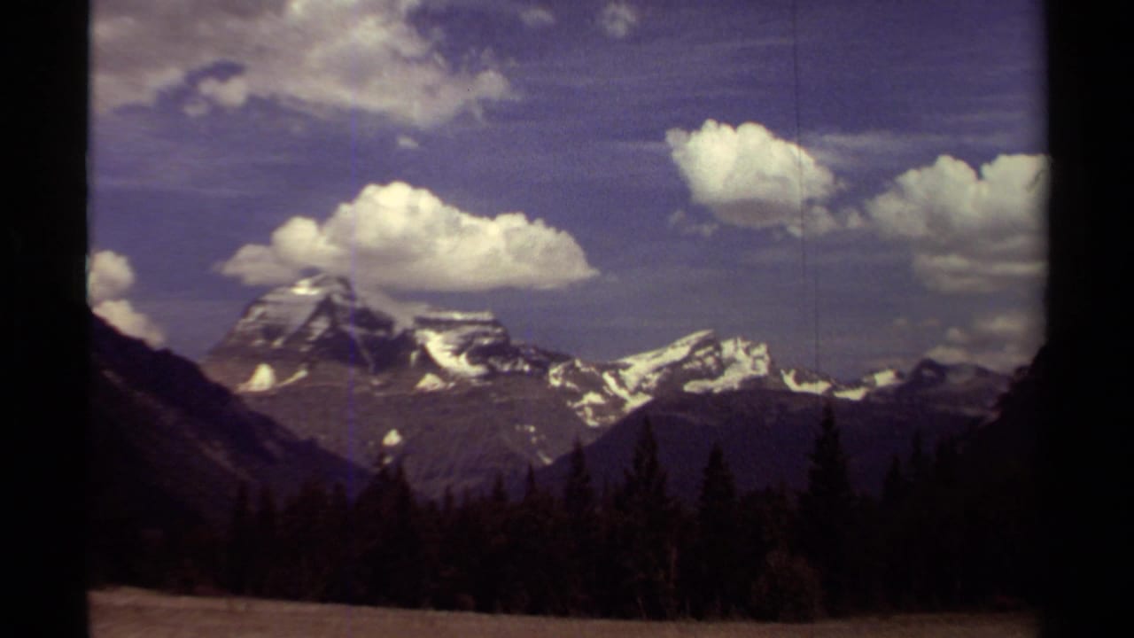 Snow-capped mountain range above a dark forested valley under scattered clouds.