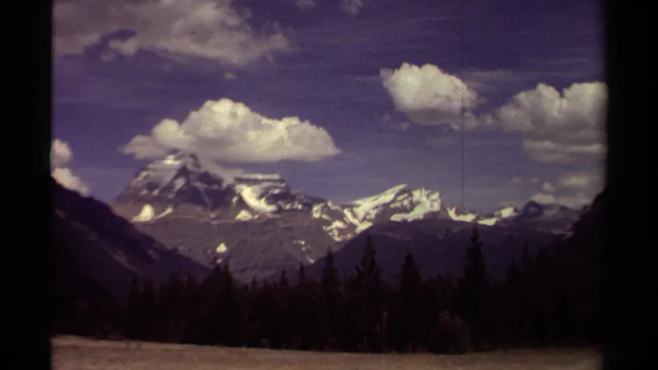 1979 British Columbia 8mm frame of Canadian Rockies scenery with a snowy ridge, dark forest foreground, and large clouds overhead.