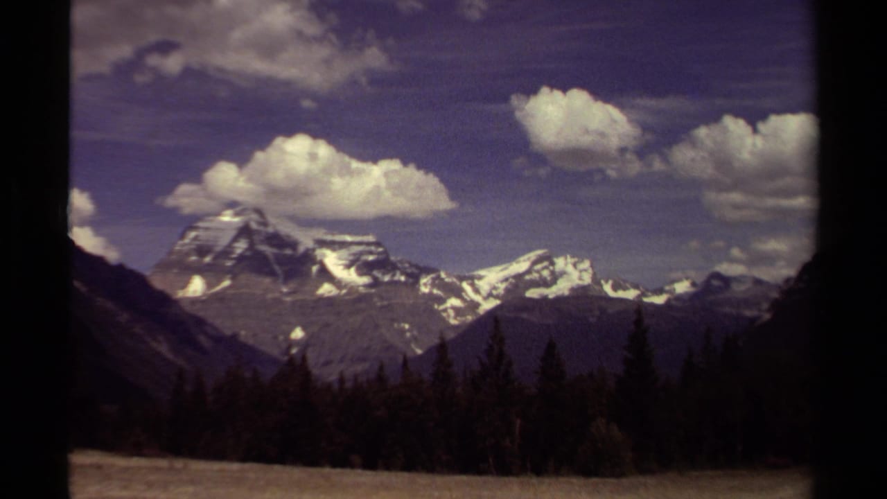 1979 8mm view in British Columbia showing distant snow-capped mountains, evergreen trees, and a partly cloudy blue sky.