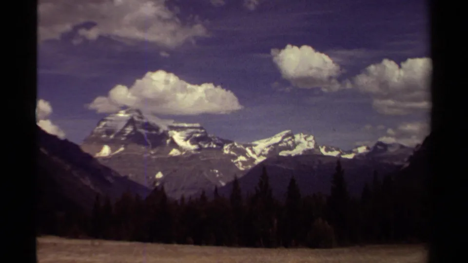 British Columbia 8mm frame from 1979 with snow-bright mountain ridges, dark forest foreground, and billowing clouds overhead.
