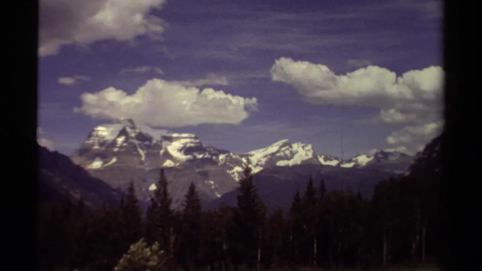Vintage 8mm British Columbia landscape frame showing snow-patched peaks, evergreen forest, and drifting clouds in a soft 1970s film palette.
