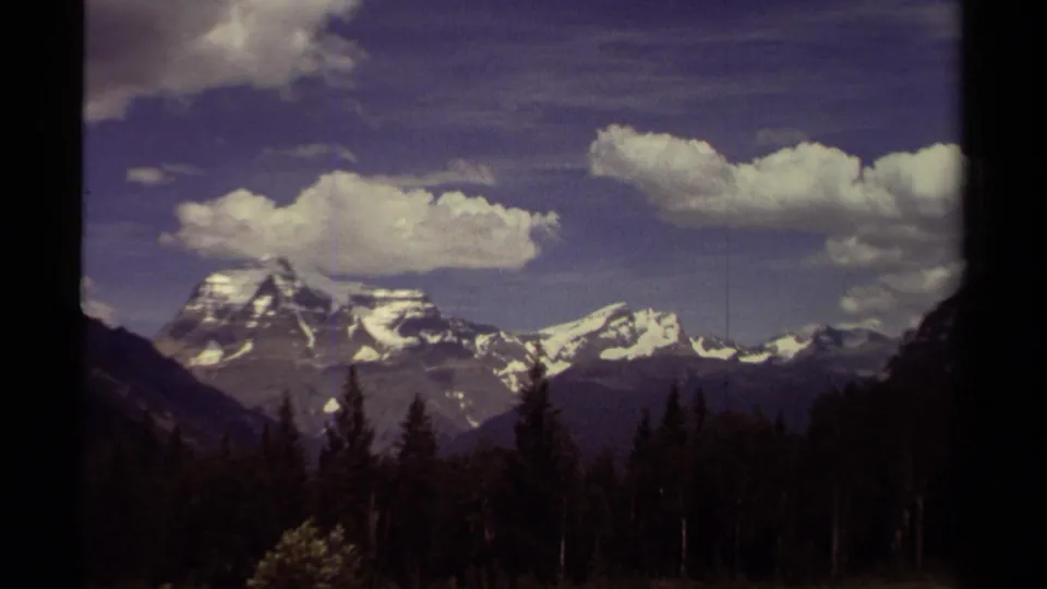 1979 British Columbia home-movie frame with distant snowy mountains, a dark evergreen foreground, and broad clouds across the sky.