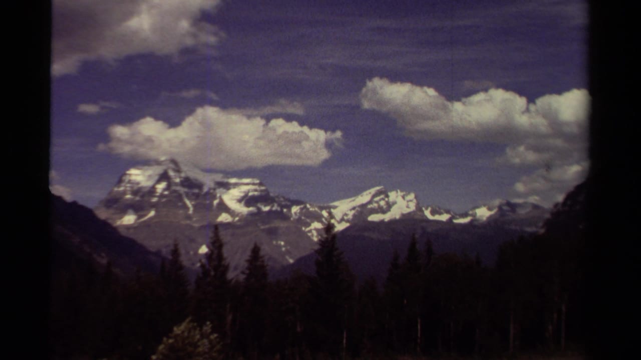 Snow-capped mountains above a dark conifer forest under scattered clouds.