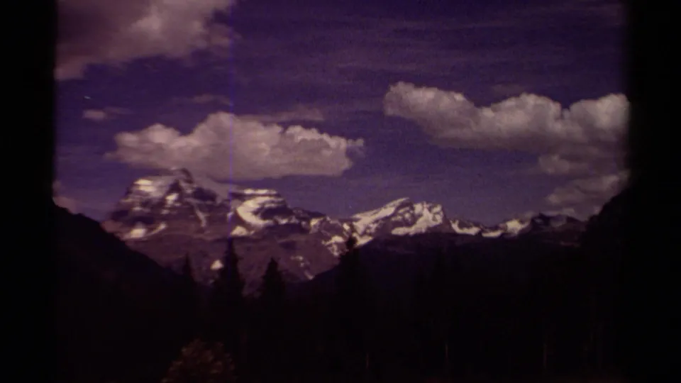 1979 British Columbia mountain view with snowy ridges, forest foreground, clouded sky, and tinted 8mm film color.