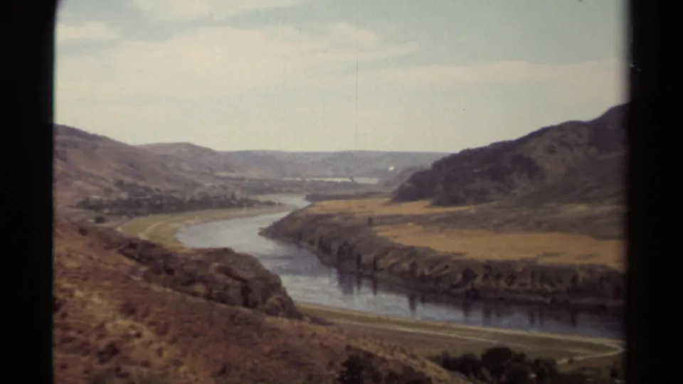 1979 British Columbia 8mm view of a winding river valley, dry hillsides, distant settlement, and pale sky.