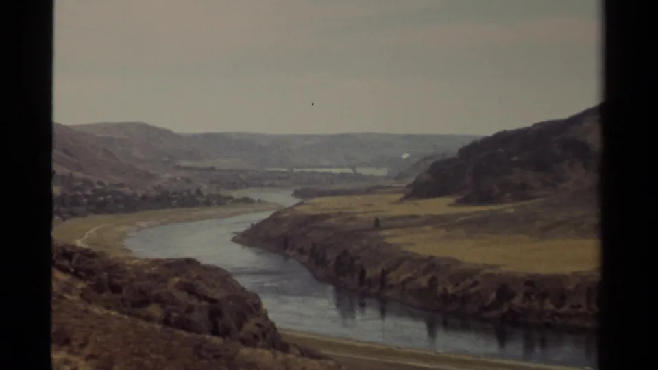 1979 British Columbia home-movie frame with the camera zooming toward a river bend and open hillside terrain.