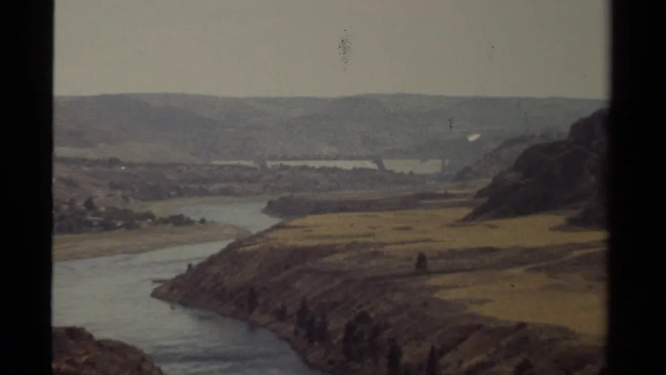1979 British Columbia 8mm frame showing the zoomed-in river valley, with muted color, dry hills, and a hazy distant ridge.