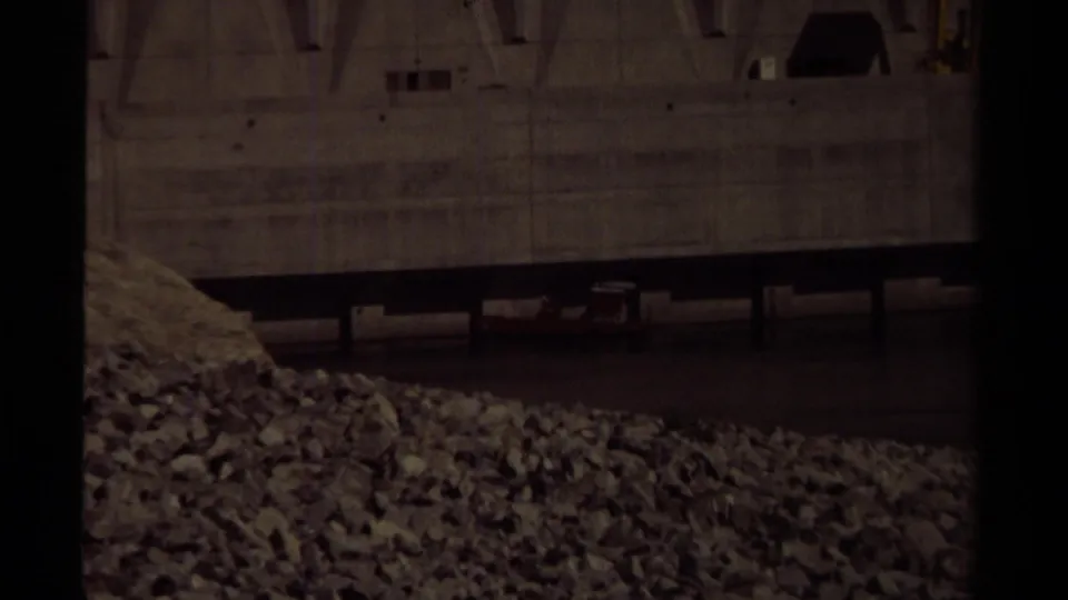 Closer 1979 8mm view of the concrete dam face in British Columbia, with gravel in the foreground and water at the base.