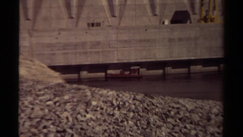 Late-1970s British Columbia home-movie frame showing the dam base, waterline, gravel foreground, and a small boat beside the concrete structure.