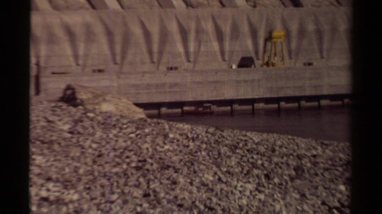 1979 British Columbia 8mm frame showing a concrete dam wall, arched spillway forms, rocky embankment, and dark river water.