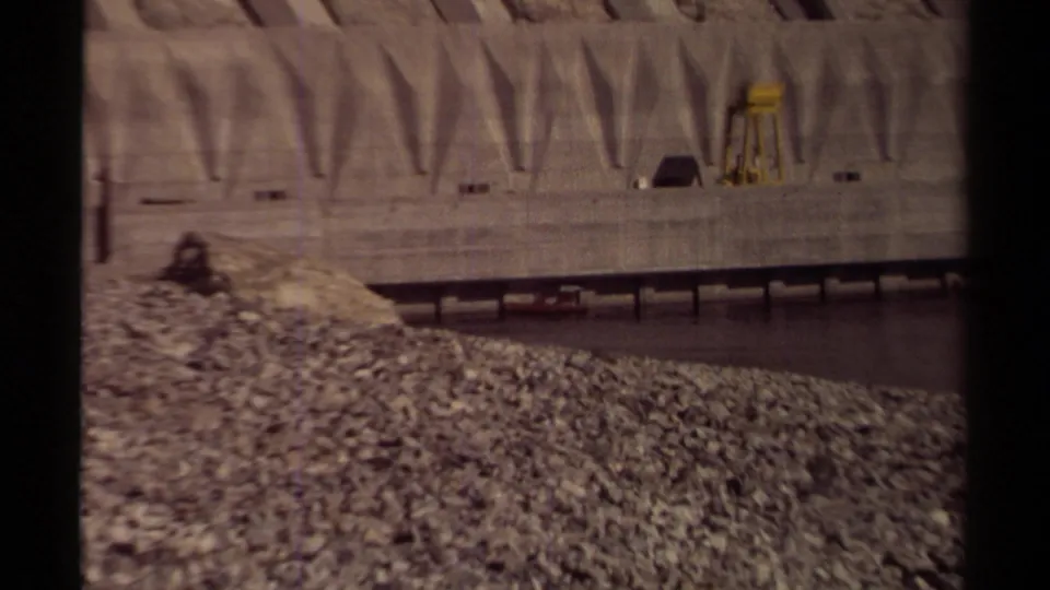 1979 British Columbia 8mm frame showing a concrete dam wall, arched spillway forms, rocky embankment, and dark river water.