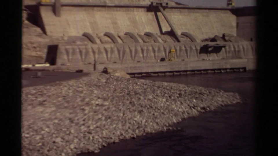1979 British Columbia home-movie frame with a wide dam view, rocky riverbank, arched concrete forms, and a small boat near the base.