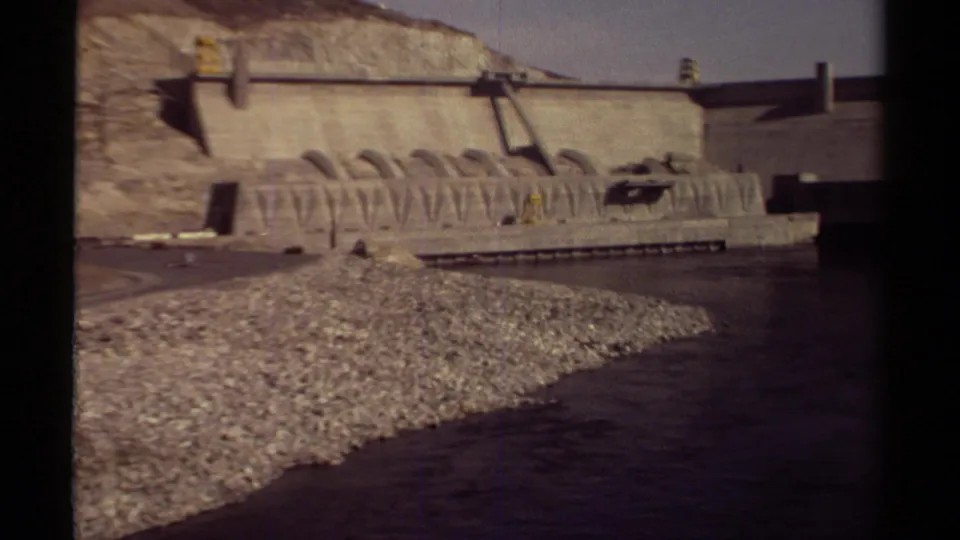 1979 British Columbia 8mm view of a large concrete dam, repeated spillway arches, rocky embankment, and broad river channel.