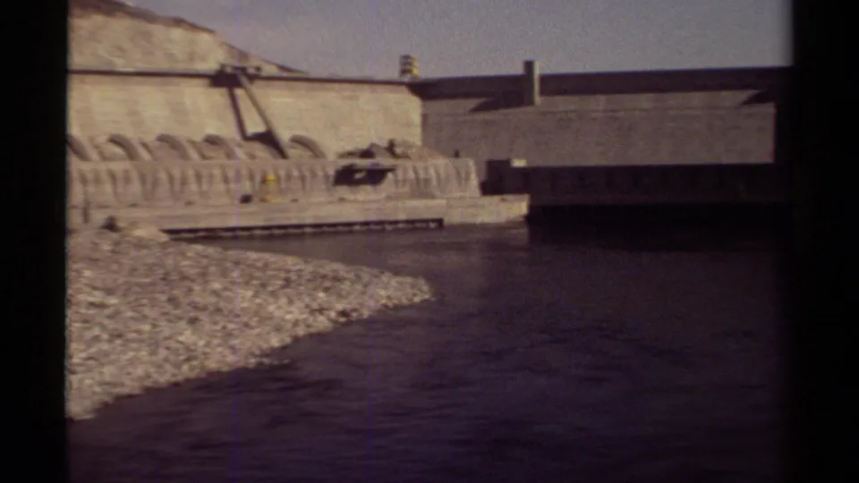 1979 British Columbia frame showing river flow beside a large concrete dam, rocky embankment, arched forms, and dark structure at right.