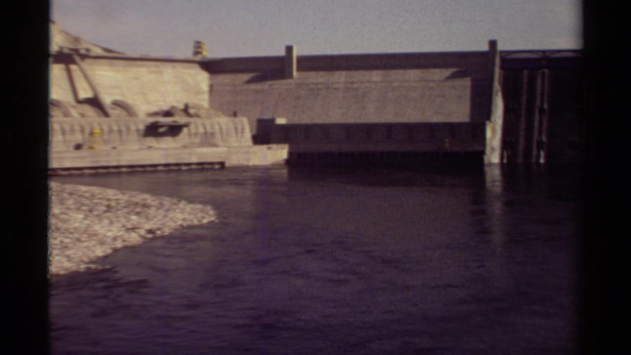 1979 British Columbia 8mm frame showing a concrete dam, dark water, sloped bank, pale sky, and black film borders.