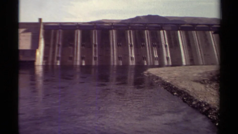 1979 British Columbia 8mm frame showing dam gates across dark water, a right-side shoreline edge, distant hills, and pale sky.