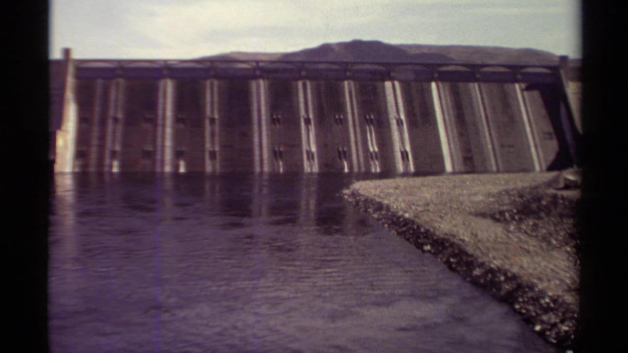 Concrete dam across calm water with gravel shoreline and distant hills.