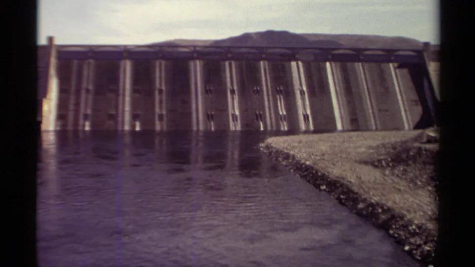 1979 British Columbia 8mm view of a concrete dam across still reservoir water, with a gravel shoreline and low hills beyond.