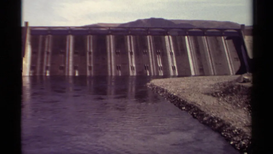 Home-movie frame from British Columbia in 1979 showing the dam face, quiet reservoir water, and gravel shoreline under a pale sky.