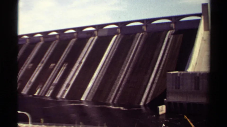 Closer 8mm view of the British Columbia dam, showing vertical spillway bays, an arched crest, dark water, and part of a concrete structure at right.