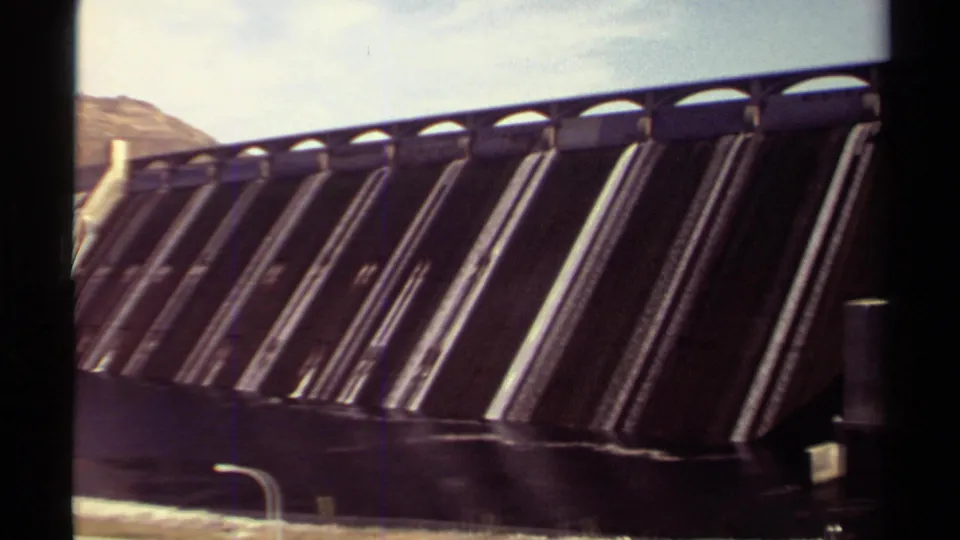 1979 British Columbia home-movie frame showing the dam's sloped concrete face, repeated spillway divisions, waterline, and hillside in the background.