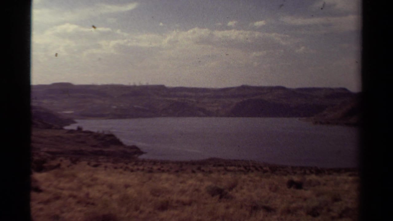 1979 8mm home-movie view of a broad lake below low hills, with a scrubby foreground and overcast sky.