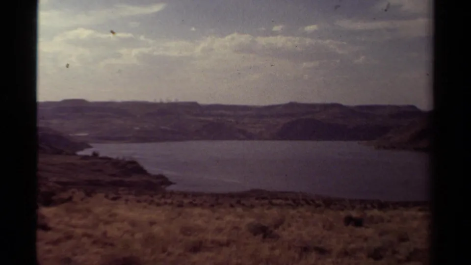 1979 8mm home-movie view of a broad lake below low hills, with a scrubby foreground and overcast sky.