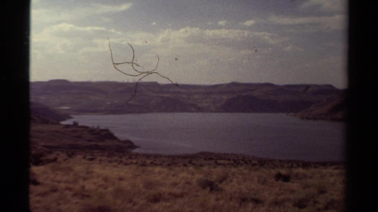 Faded 8mm frame of the lake landscape, showing surface scratches, cloudy sky, and distant hills.
