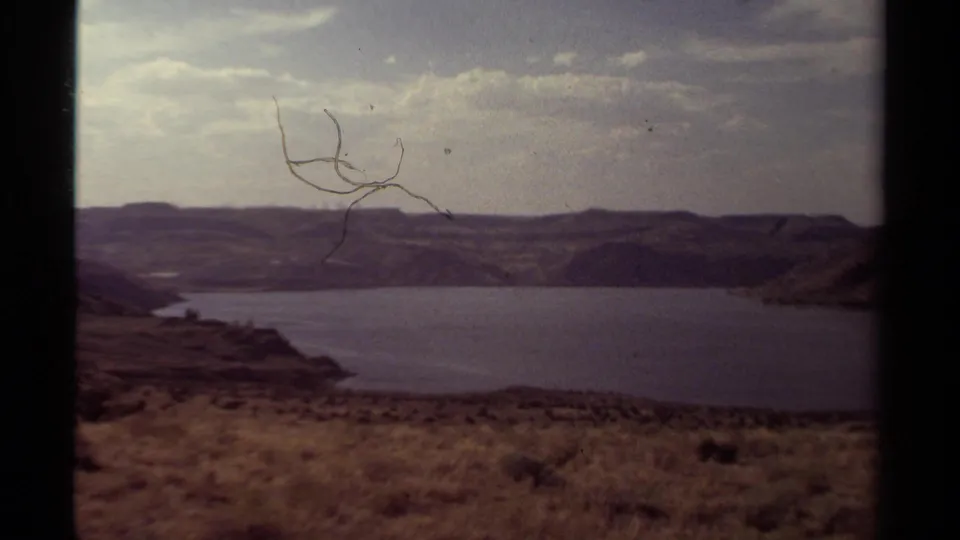 Faded 8mm frame of the lake landscape, showing surface scratches, cloudy sky, and distant hills.