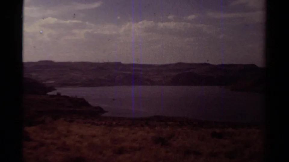 Underexposed 8mm view of the reservoir landscape, with hills, water, dry brush, and blue vertical film streaks.